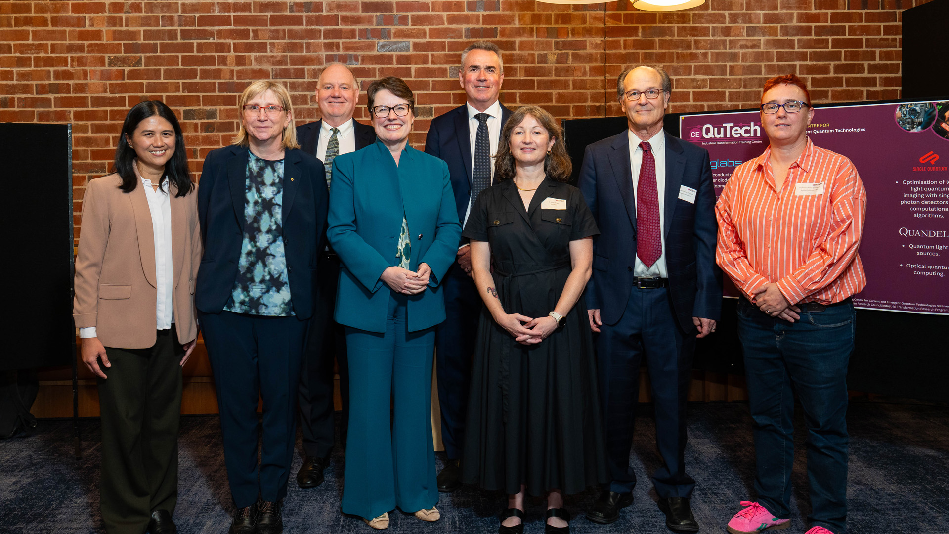 Group of professional people posing in front of a brick style wall for an event opening.